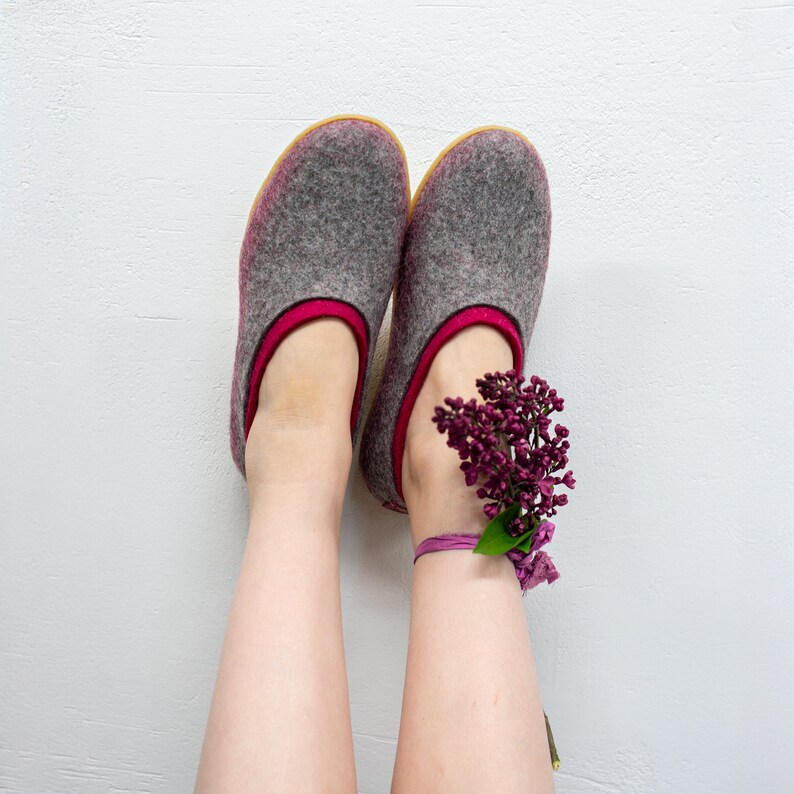 K&ouml;nnte beinhalten: Ein Paar graue Filzpantoffeln mit rosa Besatz. Die Pantoffeln werden von einer Person mit nackten Beinen getragen. Ein Strau&szlig; lila Blumen ist um ein Kn&ouml;chel gebunden.