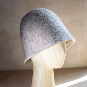 May include: A light grey felted bucket hat with a rounded crown and a short, slightly flared brim. The hat features a textured, mottled surface with subtle variations in colour, displaying a soft, dense material construction throughout.
