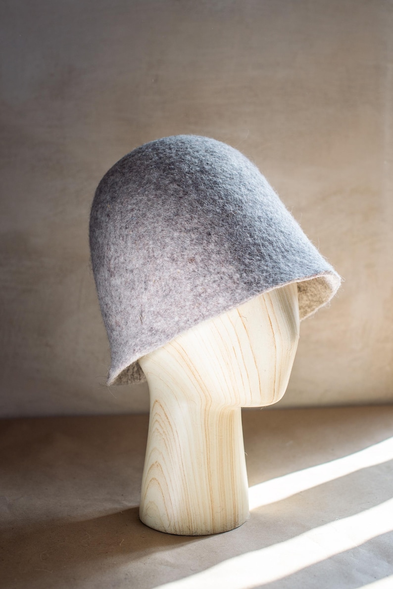 May include: A light grey felted bucket hat with a rounded crown and a short, slightly flared brim. The hat features a textured, mottled surface with subtle variations in colour, displaying a soft, dense material construction throughout.