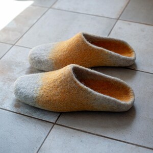 May include: A pair of felted wool slippers. The slippers have a gray toe that fades into a yellow-orange color. The interior of the slippers is a darker orange. The slippers are sitting on a tiled floor.