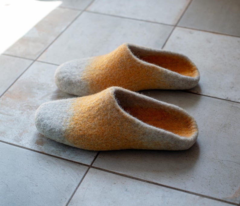 May include: A pair of felted wool slippers. The slippers have a gray toe that fades into a yellow-orange color. The interior of the slippers is a darker orange. The slippers are sitting on a tiled floor.
