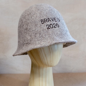May include: A light grey felt bucket hat featuring the dark brown embroidered text BRAVES 2026 on the front. The hat has a rounded crown and a short, slightly flared brim, displaying a textured, mottled appearance throughout the material.