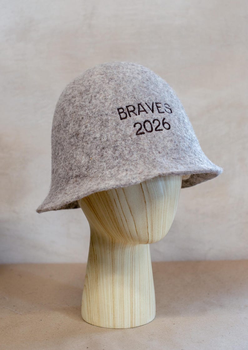 May include: A light grey felt bucket hat featuring the dark brown embroidered text BRAVES 2026 on the front. The hat has a rounded crown and a short, slightly flared brim, displaying a textured, mottled appearance throughout the material.