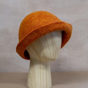 May include: A vibrant orange felted bucket hat with a rounded crown and a slightly downturned brim. The hat features a textured, mottled surface with subtle variations in orange and yellow tones, showcasing a dense, soft wool-like material.