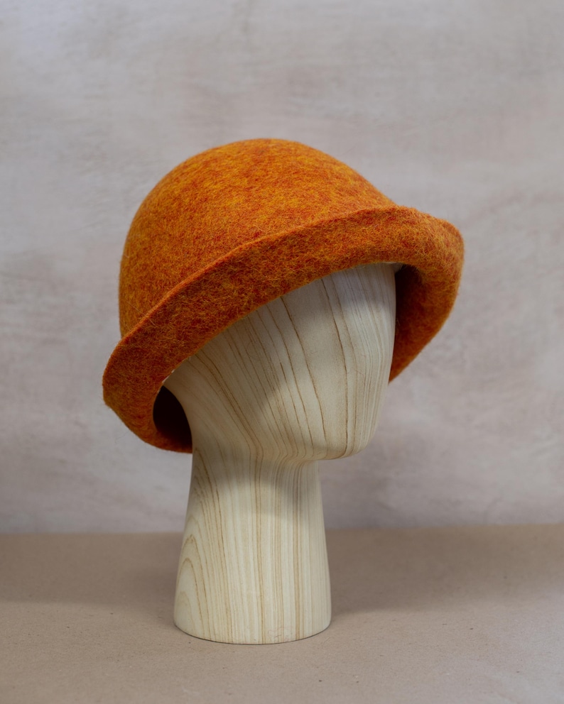 May include: A vibrant orange felted bucket hat with a rounded crown and a slightly downturned brim. The hat features a textured, mottled surface with subtle variations in orange and yellow tones, showcasing a dense, soft wool-like material.