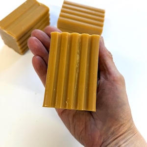 以下が含まれることがあります： 3本の茶色の石鹸。石鹸は溝のある質感で、手の中に置かれています