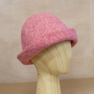 May include: A pink felted wool hat with a rounded crown and a slightly upturned brim. The hat features a mottled texture with varying shades of pink and white fibres throughout the material, creating a soft, dense appearance on the dome-shaped head.