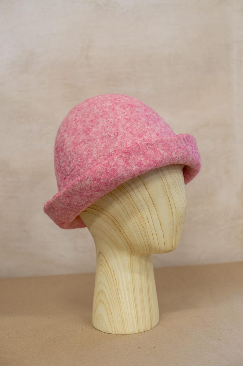 May include: A pink felted wool hat with a rounded crown and a slightly upturned brim. The hat features a mottled texture with varying shades of pink and white fibres throughout the material, creating a soft, dense appearance on the dome-shaped head.