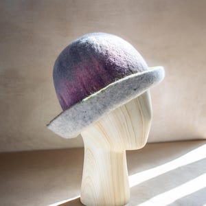 May include: A felt cloche hat in shades of purple and gray, displayed on a wooden head form. The hat has a rounded crown and a wide, downturned brim. The brim is gray, and the crown transitions from purple to gray.
