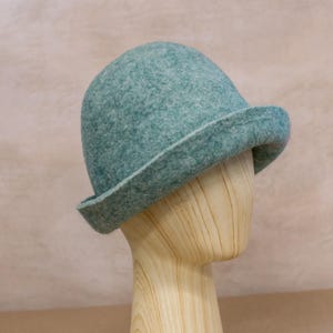 May include: A teal felted wool hat with a rounded crown and a slightly upturned brim. The hat features a textured, mottled surface with subtle variations in colour, showcasing a dense, soft material construction that maintains a structured shape.