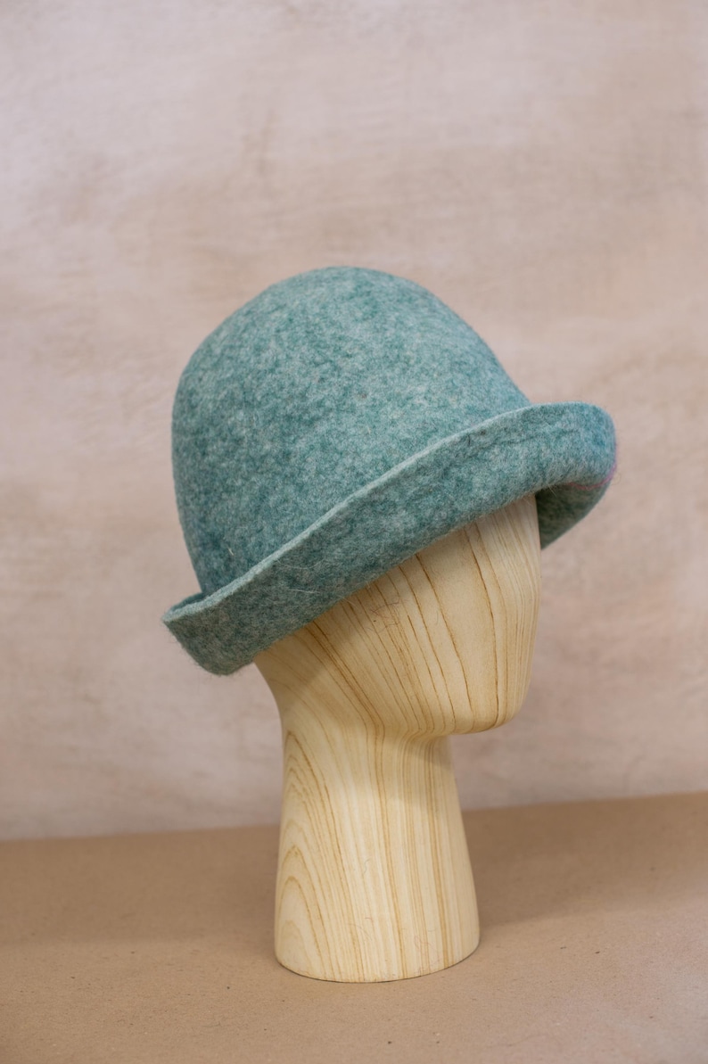 May include: A teal felted wool hat with a rounded crown and a slightly upturned brim. The hat features a textured, mottled surface with subtle variations in colour, showcasing a dense, soft material construction that maintains a structured shape.