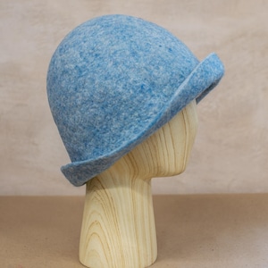 May include: A light blue felted wool cloche hat with a rounded crown and a short, slightly upturned brim. The material has a mottled texture with variations of blue and white fibres throughout the surface, creating a soft, heathered appearance.