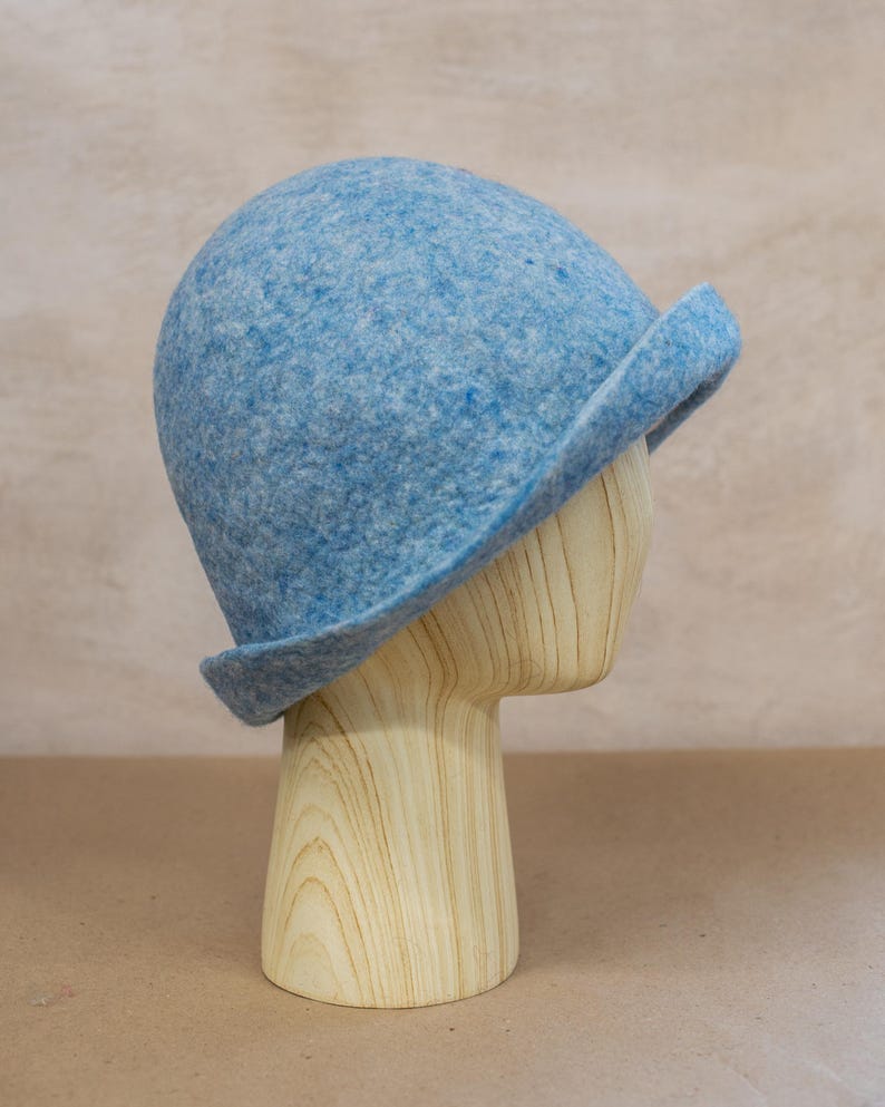 May include: A light blue felted wool cloche hat with a rounded crown and a short, slightly upturned brim. The material has a mottled texture with variations of blue and white fibres throughout the surface, creating a soft, heathered appearance.