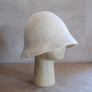 May include: A solid off-white felted hat with a rounded crown and a slightly flared, asymmetrical brim. The hat features a soft, textured surface and a simple, unadorned design, showcasing the natural appearance of the felted material throughout.