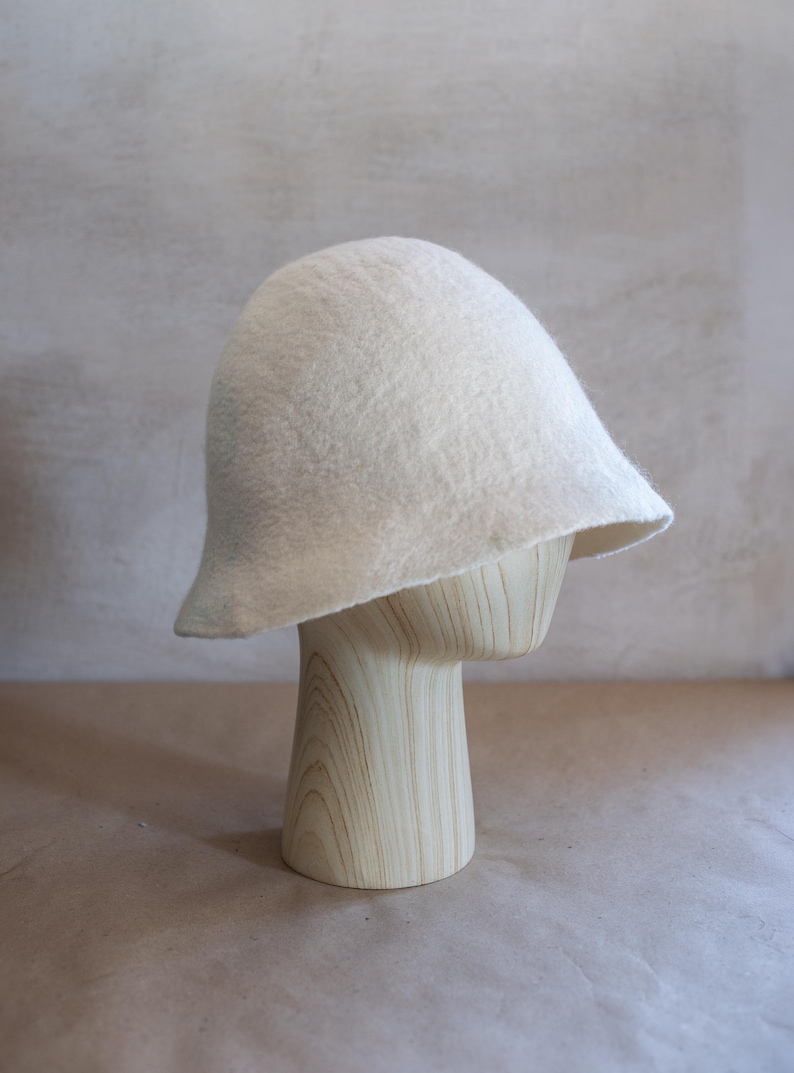 May include: A solid off-white felted hat with a rounded crown and a slightly flared, asymmetrical brim. The hat features a soft, textured surface and a simple, unadorned design, showcasing the natural appearance of the felted material throughout.