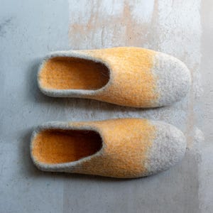 May include: A pair of handmade felt slippers. The slippers are a blend of yellow and gray, with the yellow color fading into gray at the toe. The slippers have an open heel and a rounded toe design.