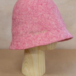 May include: A pink felted bucket hat with a mottled texture and the name Gabriel embroidered in light-coloured thread on the side. The hat features a rounded crown and a short, slightly flared brim, showcasing a soft, dense material construction.