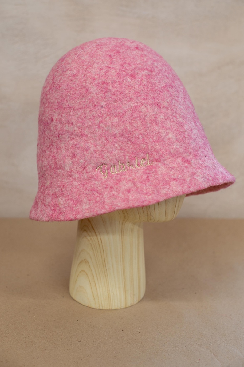 May include: A pink felted bucket hat with a mottled texture and the name Gabriel embroidered in light-coloured thread on the side. The hat features a rounded crown and a short, slightly flared brim, showcasing a soft, dense material construction.