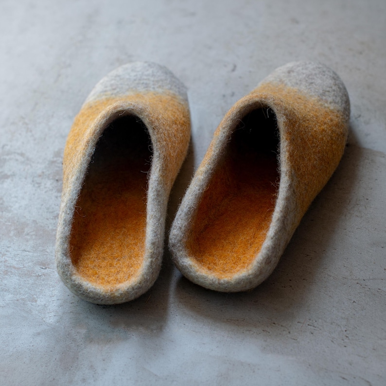 May include: A pair of felted wool slippers. The slippers have a gradient design, transitioning from gray at the toe to yellow-orange in the middle and inside. The edges are gray. The slippers are designed for indoor use.