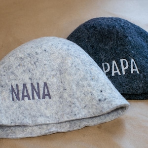 May include: Two felted wool hats, one light grey with the word NANA printed in dark grey, and one dark charcoal grey with the word PAPA printed in white. Both hats feature a rounded, dome-shaped crown and a simple, clean-edged brim design.