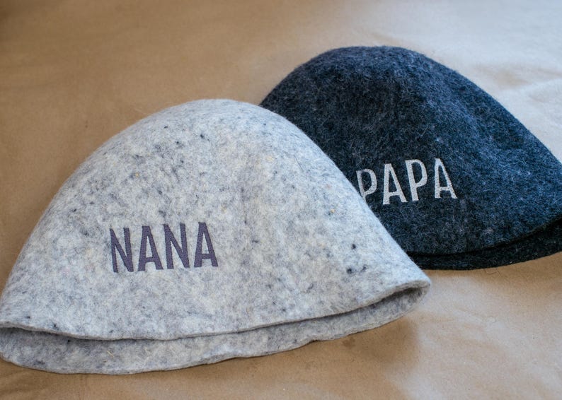 May include: Two felted wool hats, one light grey with the word NANA printed in dark grey, and one dark charcoal grey with the word PAPA printed in white. Both hats feature a rounded, dome-shaped crown and a simple, clean-edged brim design.