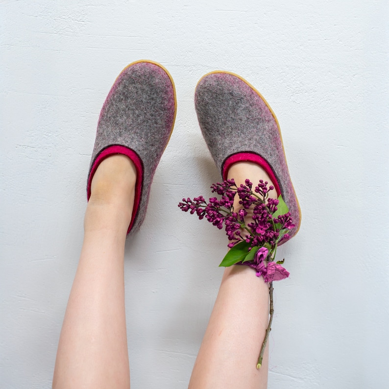 K&ouml;nnte beinhalten: Ein Paar graue Filzpantoffeln mit rosa Besatz, getragen von einer Person mit einem Strau&szlig; lila und rosa Blumen, die in die Oberseite eines Pantoffels gesteckt sind.