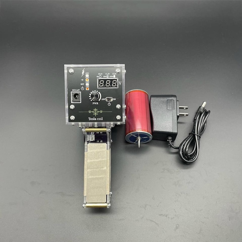 以下が含まれることがあります： 透明なアクリル製のケース、赤いコイル、電源、および「8.88 V」を表示するデジタルディスプレイを備えた黒いテスラコイルキット。キットには、電源スイッチ、ノブ、および「テスラコイル」というラベルが含まれています。