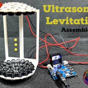 以下が含まれることがあります： 白の3Dプリントフレームに、上部と下部に黒い超音波センサーの円形アレイが配置されています。フレームは、ワイヤーで青い回路基板に接続されています。テキスト「Ultrasonic Levitation Assembled!!!」は、黒い背景に黄色で書かれています。