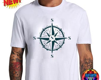 May include: A white short-sleeved t-shirt featuring a dark green compass rose graphic centered on the chest. The compass design includes the letter N in the center and the letter S at each of the four cardinal points. A yellow starburst graphic with the text NEW is in the top left corner.