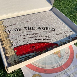 May include: A white jigsaw puzzle of a world map in a box. The puzzle is titled "Map of the World" and is labeled "Practically Correct" with the creator's name and location. The puzzle is partially assembled and shows a red ruler with Roman numerals.