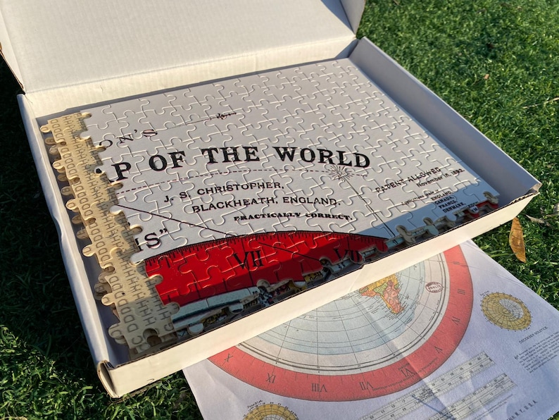 May include: A white jigsaw puzzle of a world map in a box. The puzzle is titled "Map of the World" and is labeled "Practically Correct" with the creator's name and location. The puzzle is partially assembled and shows a red ruler with Roman numerals.