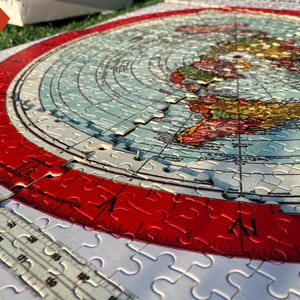 May include: A flat earth jigsaw puzzle with a red border and a map of the world. The puzzle is partially assembled and shows the continents of North America, South America, Europe, Africa, and Asia. The puzzle is made by Flat Earth Products.