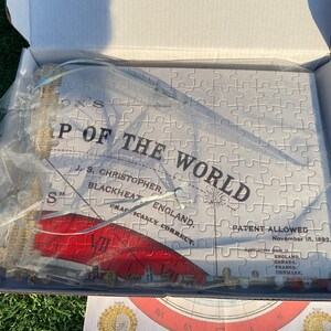 May include: A white jigsaw puzzle in a box with a blue and white ribbon. The puzzle is of a map of the world with the text "P of the World" and "Patent Allowed November 15, 1992".