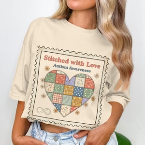 May include: Cream-colored t-shirt featuring a heart-shaped patchwork design with the text "Stitched with Love Autism Awareness." The design is framed within a postage stamp border, with small floral and infinity symbol details.