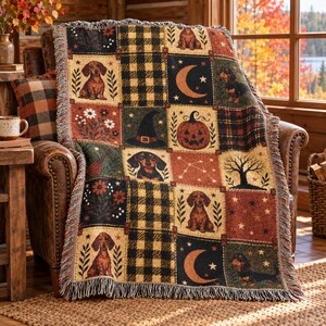 May include: A woven throw blanket featuring a patchwork design with dachshund dogs, pumpkins, witch hats, crescent moons, stars, and trees. The pattern includes black and tan buffalo plaid squares in a warm autumn color palette with fringe edges.