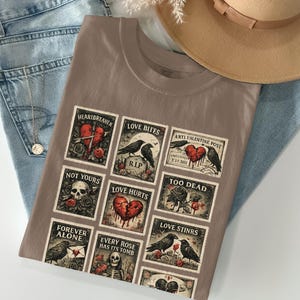 May include: A taupe t-shirt featuring a collage of anti-Valentine's Day themed postage stamp designs. The stamps depict broken hearts, crows, roses, and skulls, with phrases like "Heartbreaker," "Love Bites," and "Forever Alone."