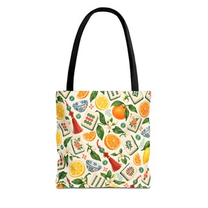 May include: A tote bag with black handles featuring a cream-coloured background patterned with orange slices, lemons, green leaves, white blossoms, blue and white patterned bowls, mahjong tiles, and red decorative tassels with green beads.