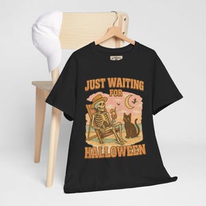 May include: Black t-shirt with a Halloween-themed graphic. The design features a skeleton in a beach chair with a drink, a black cat, and the text "JUST WAITING FOR HALLOWEEN". A white baseball cap hangs on a wooden chair.