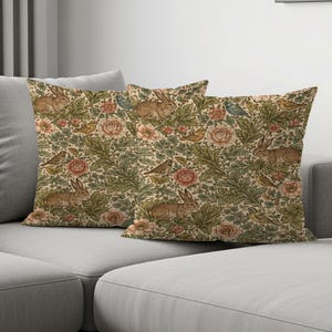 Può includere: Due cuscini decorativi con un motivo ripetuto di conigli, uccelli e fiori nei toni del beige, verde e rosa. I cuscini sono su un divano grigio. Il design ha un'estetica botanica vintage.