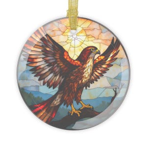 May include: Round glass ornament featuring a stained-glass style design of a hawk with outstretched wings. The hawk is perched on a branch against a backdrop of a sunset and mountains. The ornament has a gold ribbon for hanging.