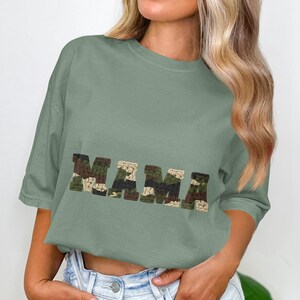 May include: Sage green t-shirt with the word "MAMA" in a camouflage pattern across the front. The shirt has short sleeves and a relaxed fit. The design is centered on the chest.