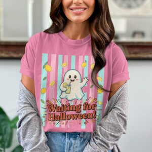Può includere: T-shirt rosa con un disegno di un fantasma dei cartoni animati che tiene in mano una bevanda, circondato da limoni e la scritta "Waiting for Halloween!". Il design presenta strisce blu e rosa. La maglietta è indossata da una persona.