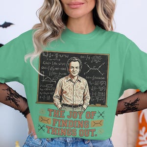May include: A light green t-shirt with a graphic of a man and equations on a chalkboard. The text "THE JOY OF FINDING THINGS OUT" is below the image. The shirt is a casual style, suitable for everyday wear.