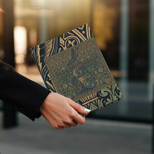 May include: A decorative book cover with a detailed illustration in shades of green, blue, and gold. The cover features an ornate design with floral and animal motifs. The book is held by a person wearing a black blazer.