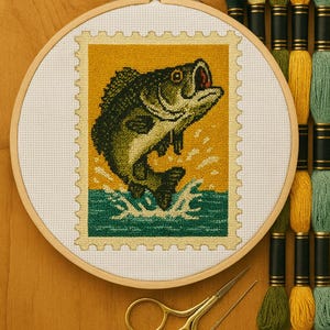 Pode incluir: Bordado em ponto cruz de um peixe saltando, semelhante a um selo postal, em um bastidor de madeira. O peixe é verde e preto sobre um fundo amarelo. Fios de bordar e tesouras douradas também são visíveis.