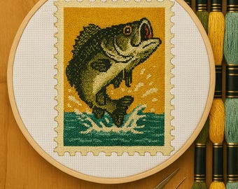May include: A cross-stitch embroidery of a leaping fish, resembling a postage stamp, set in a wooden hoop. The design features a fish in shades of green and black against a yellow background, with green water. Gold scissors and thread spools are also visible.