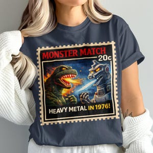 May include: Navy blue t-shirt featuring a vintage-style graphic of a monster match. The design includes two battling monsters, flames, and the text "MONSTER MATCH 20c HEAVY METAL IN 1976!" in a postage stamp style.