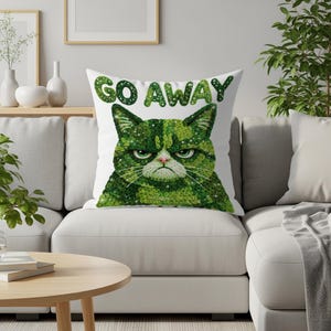 Puede incluir: Cojín cuadrado blanco con un diseño de gato verde gruñón y las palabras "GO AWAY" en texto verde. El gato tiene una expresión severa y está hecho de un patrón texturizado. El cojín está sobre un sofá blanco.