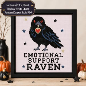 May include: A cross-stitch pattern in a black frame featuring a black raven with red eyes holding a red heart pendant in its beak. The text reads "EMOTIONAL SUPPORT RAVEN" in black letters, surrounded by small blue and yellow stars on a light fabric.