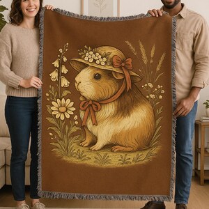 Peut inclure: Plaid marron avec une illustration de cochon d'Inde portant un chapeau fleuri et un ruban. Le motif comprend des fleurs et du blé. La couverture a une bordure à franges. La couverture est tenue par deux personnes.